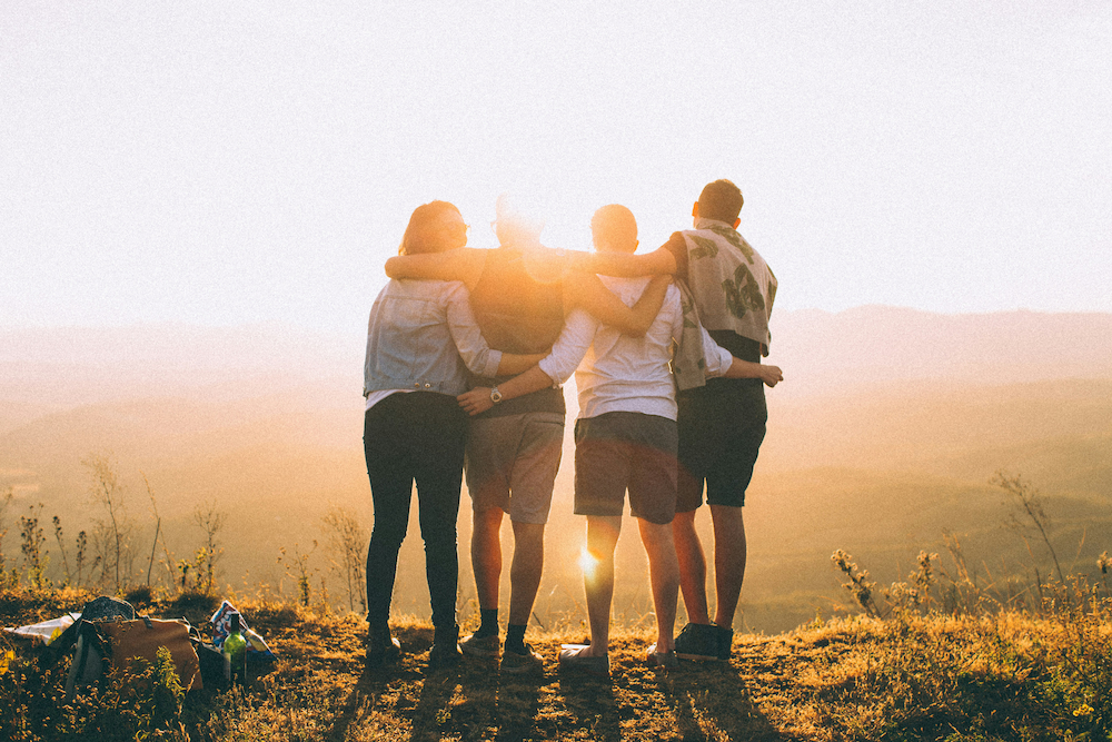 Four people standing together with arms around each other, silhouetted against a warm golden sunset over rolling hills