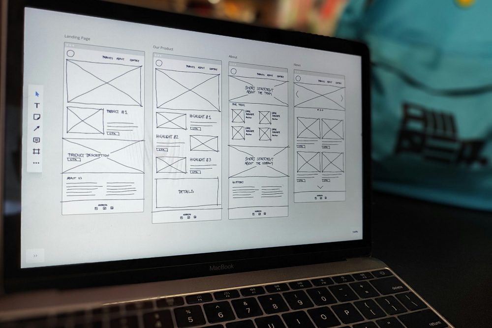 A MacBook laptop displaying multiple hand-drawn website wireframe layouts including landing page, product page, and about page sketches