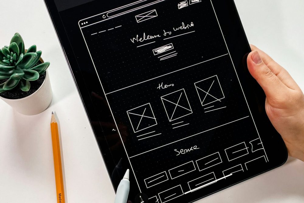 Designer holding a tablet displaying a hand-drawn website wireframe layout with pencil and stylus nearby