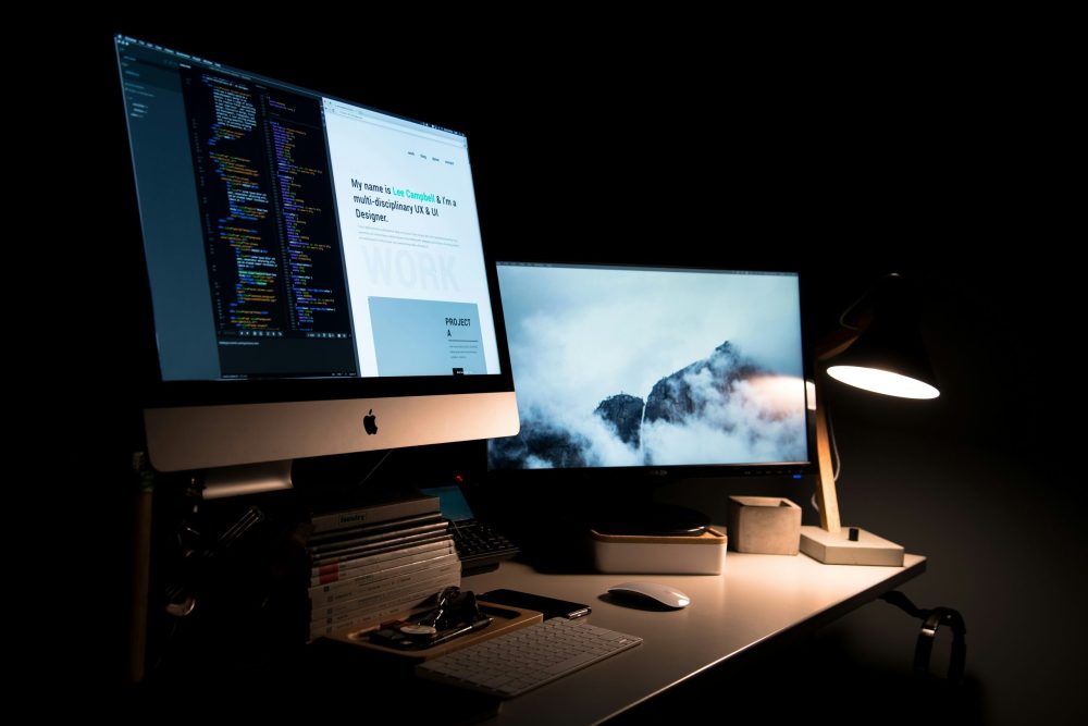 A dark web designer's desk setup with dual monitors displaying code and a portfolio website, illuminated by a warm desk lamp