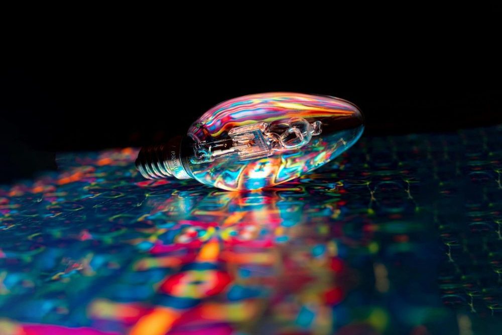 A glass lightbulb lying on its side reflecting vivid rainbow colours against a dark background