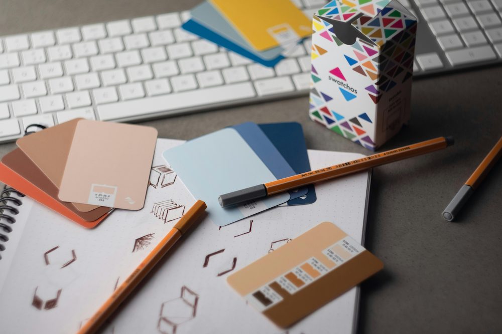 A flatlay of branding materials including colour swatches, a sketchbook with logo drawings, pens, and a colourful geometric packaging box on a desk