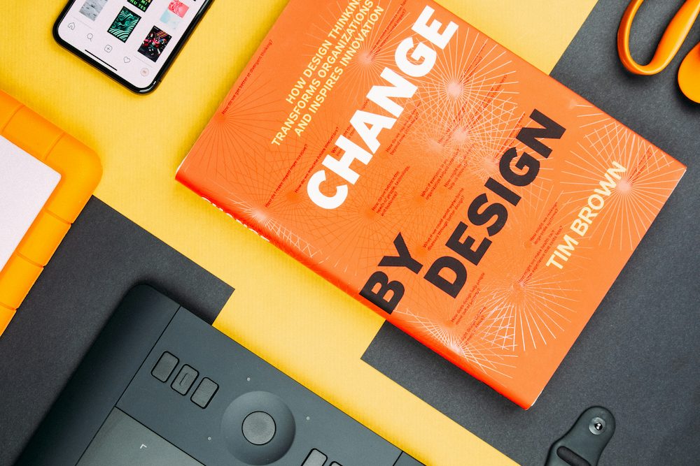 An overhead flatlay of creative design tools including a drawing tablet, smartphone, scissors, pen, and a book titled Change by Design on a yellow and black background