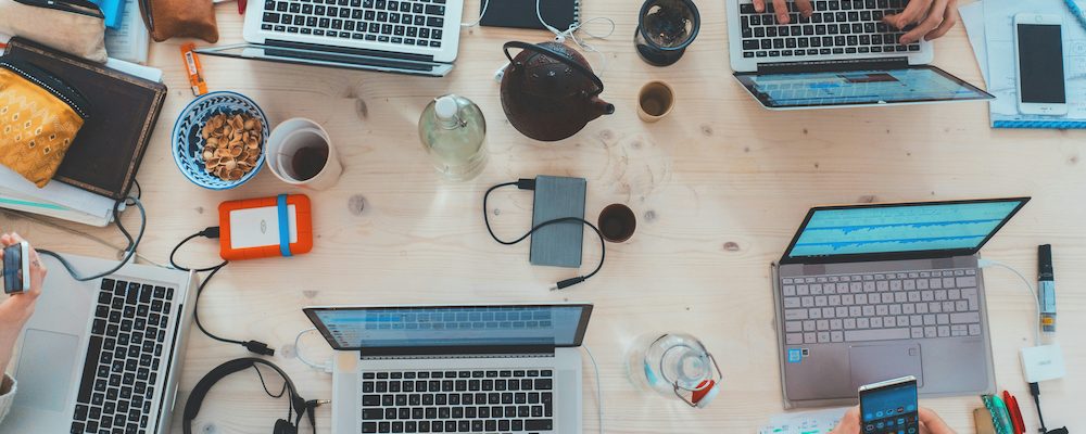 An overhead view of a busy collaborative workspace with multiple laptops, tablets, phones, headphones, and coffee on a light wood table