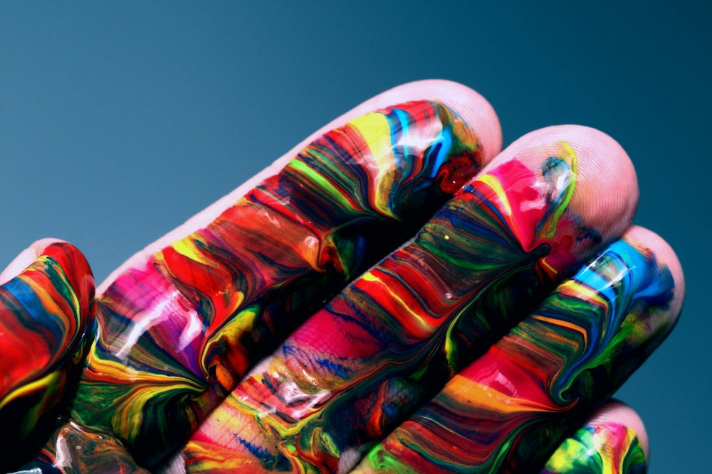 A hand covered in swirls of vibrant multicoloured paint against a teal background